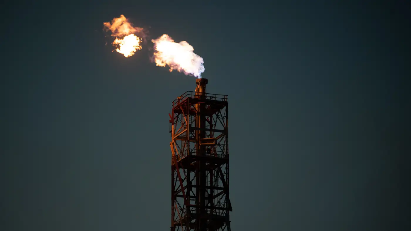 A flare stack burning against a dark dusk sky, the flame the only bright point in the frame.