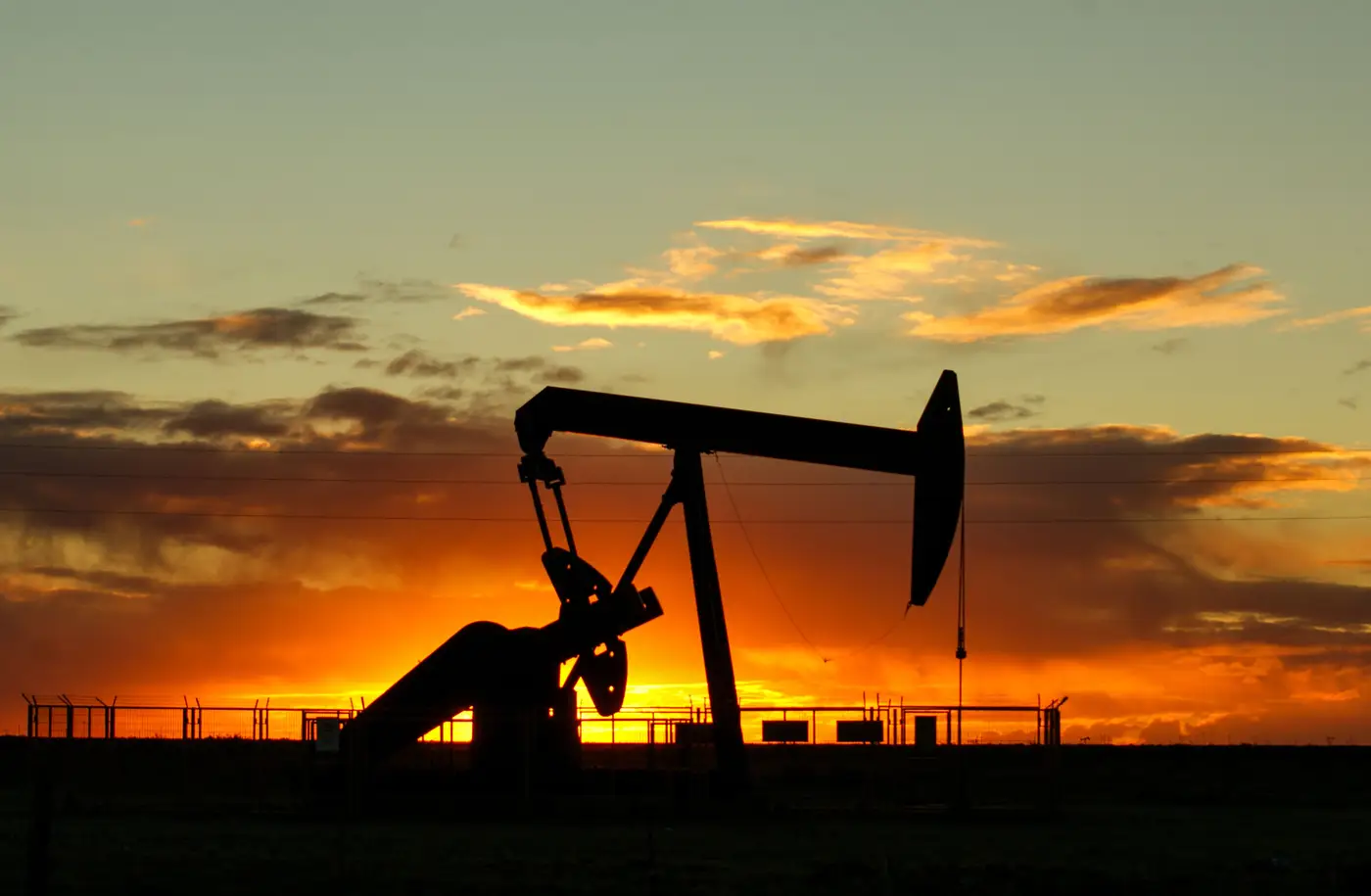 Silhouette of an oil pump jack against a vivid orange sunset sky.
